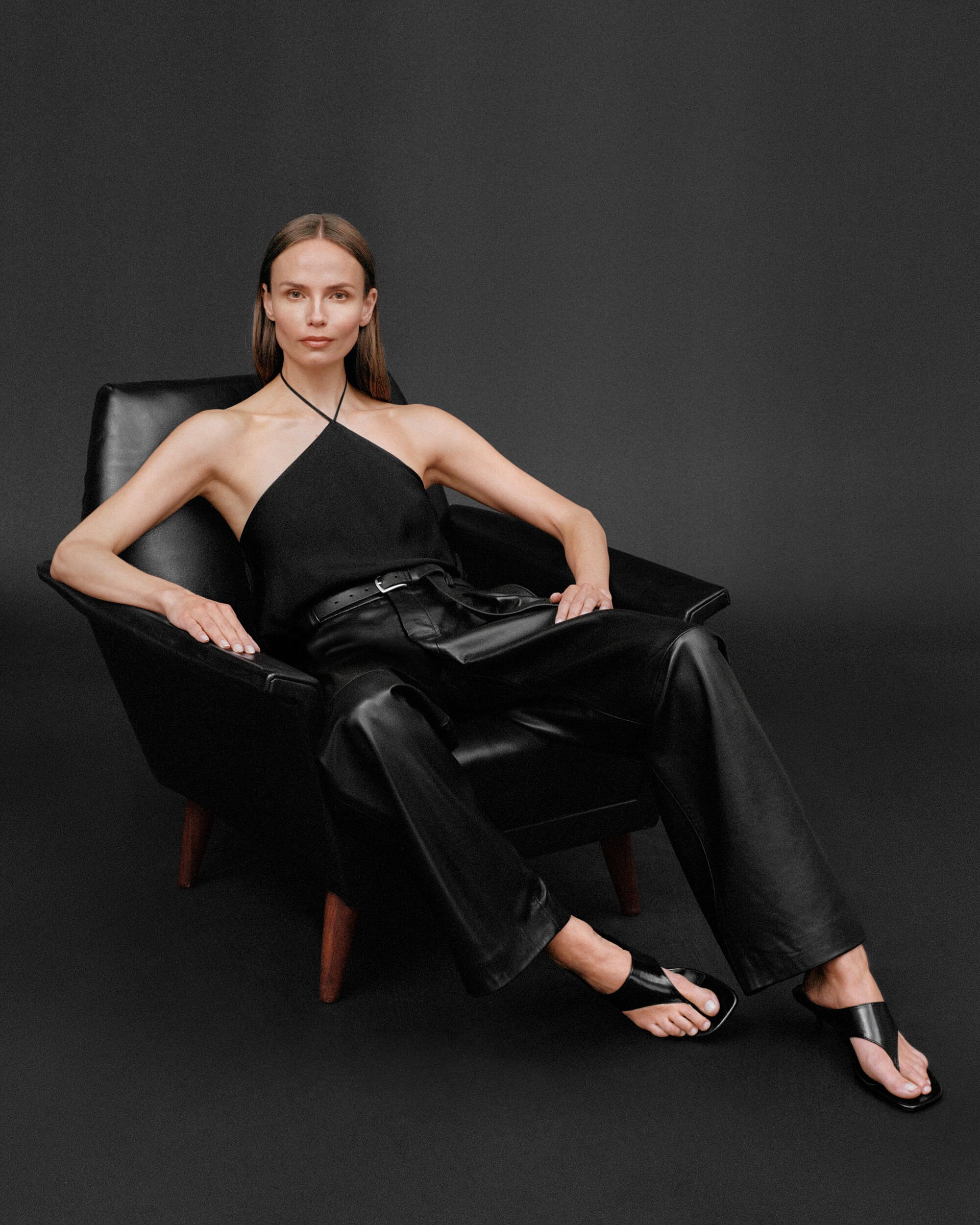 Woman in black halter top and leather trousers seated in a black armchair against a dark backdrop