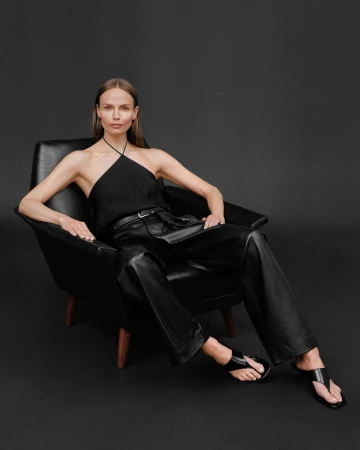 Woman in black halter top and leather trousers seated in a black armchair against a dark backdrop