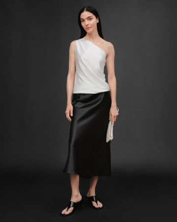 Model in a white one-shoulder top and black skirt holding a clutch against a dark grey seamless studio background.