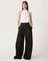 A female model standing against a white seamless background wearing a white halter top and high waisted black wide leg trousers.