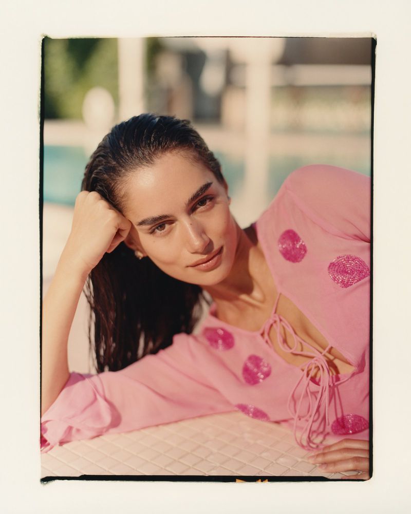 Brunette woman with wet hair poses in pink top with sequin polka dots, resting on a patterned surface with a blurred outdoor background.