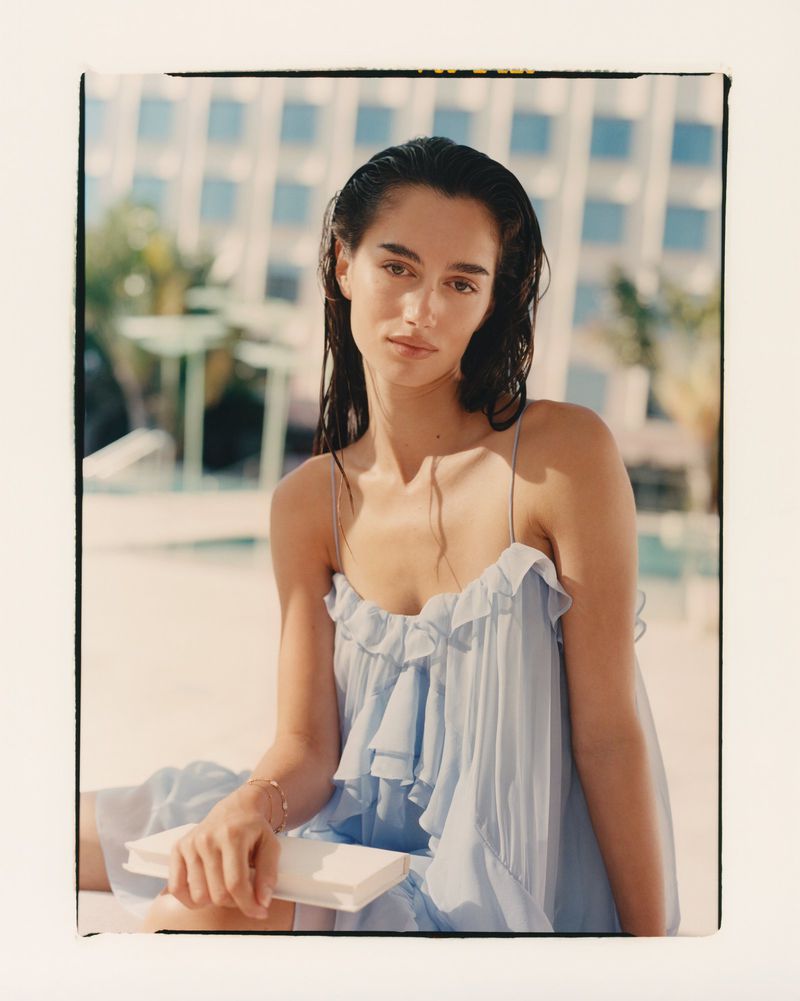 Woman in ruffled dress sits with a book outdoors by a pool, with a modern building and greenery in the background, in a sunlit scene.