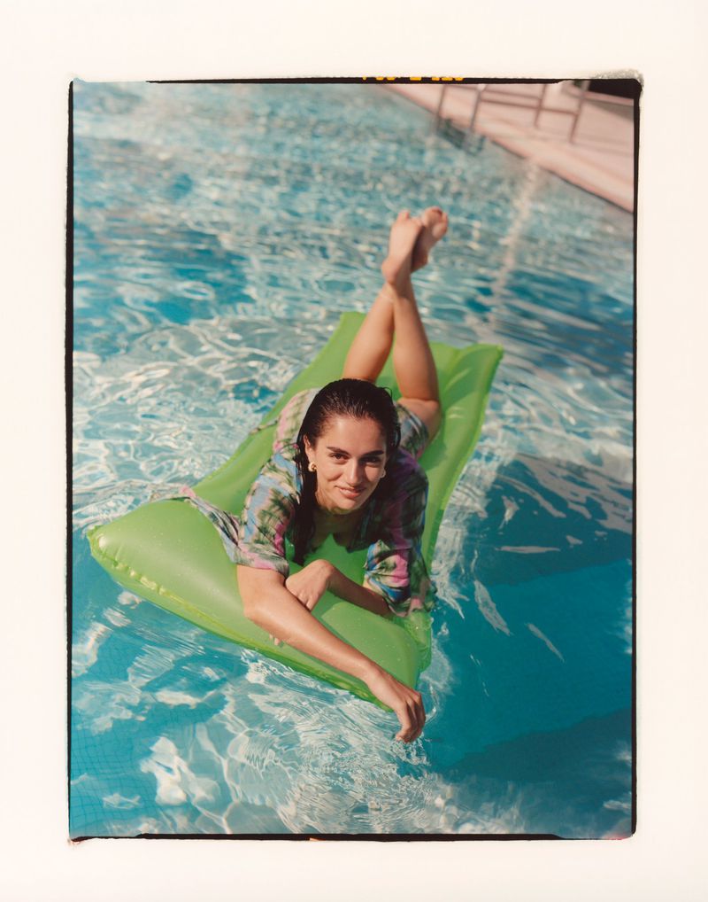 A woman in a floral shirt is lying on a lime green inflatable raft in a blue swimming pool with her feet raised slightly.