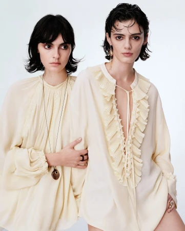 Two women posing together wearing cream colored ruffled blouses against a plain light background