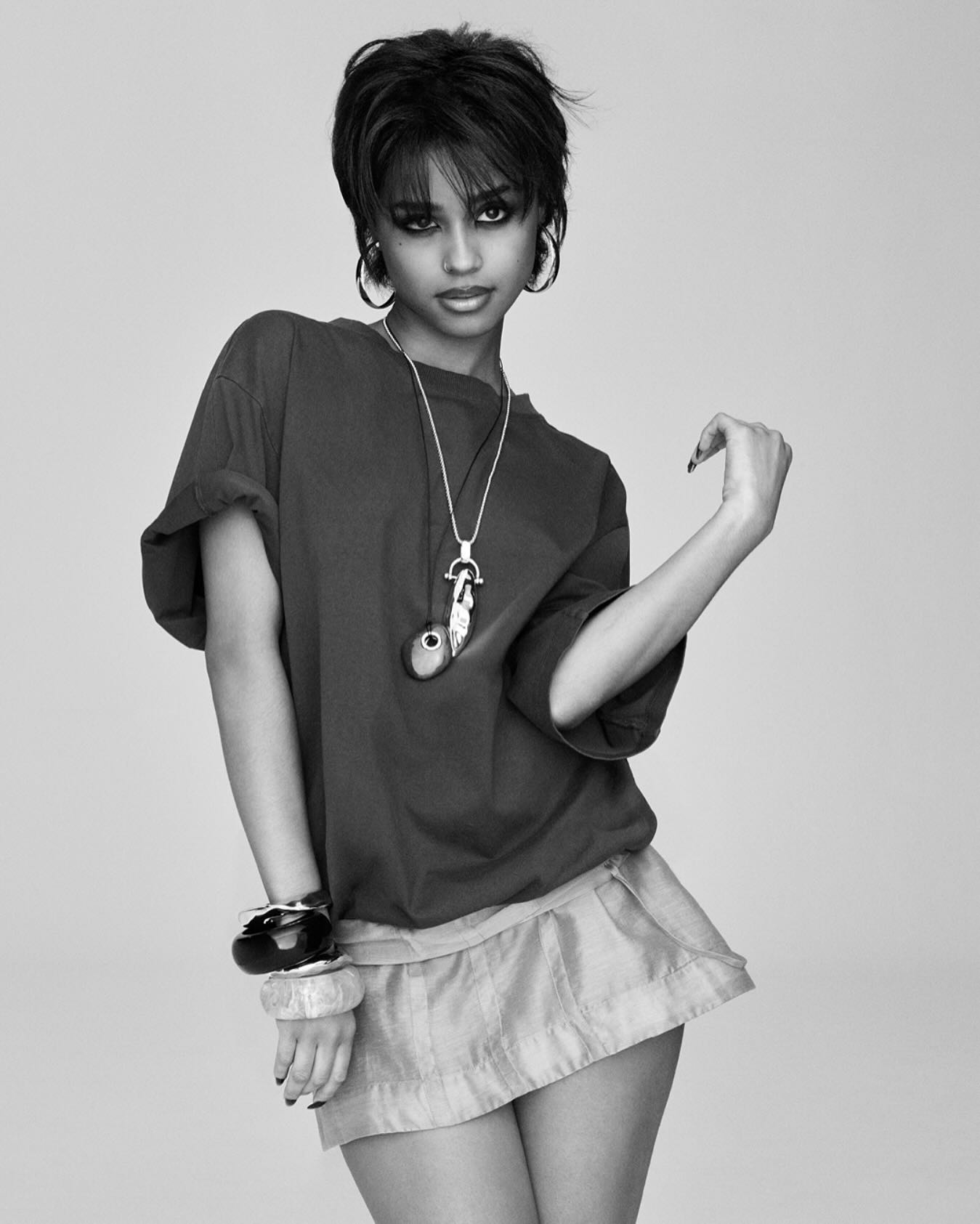 Model in dark oversized t-shirt and light mini skirt wearing layered necklaces and bracelets against a grey seamless background
