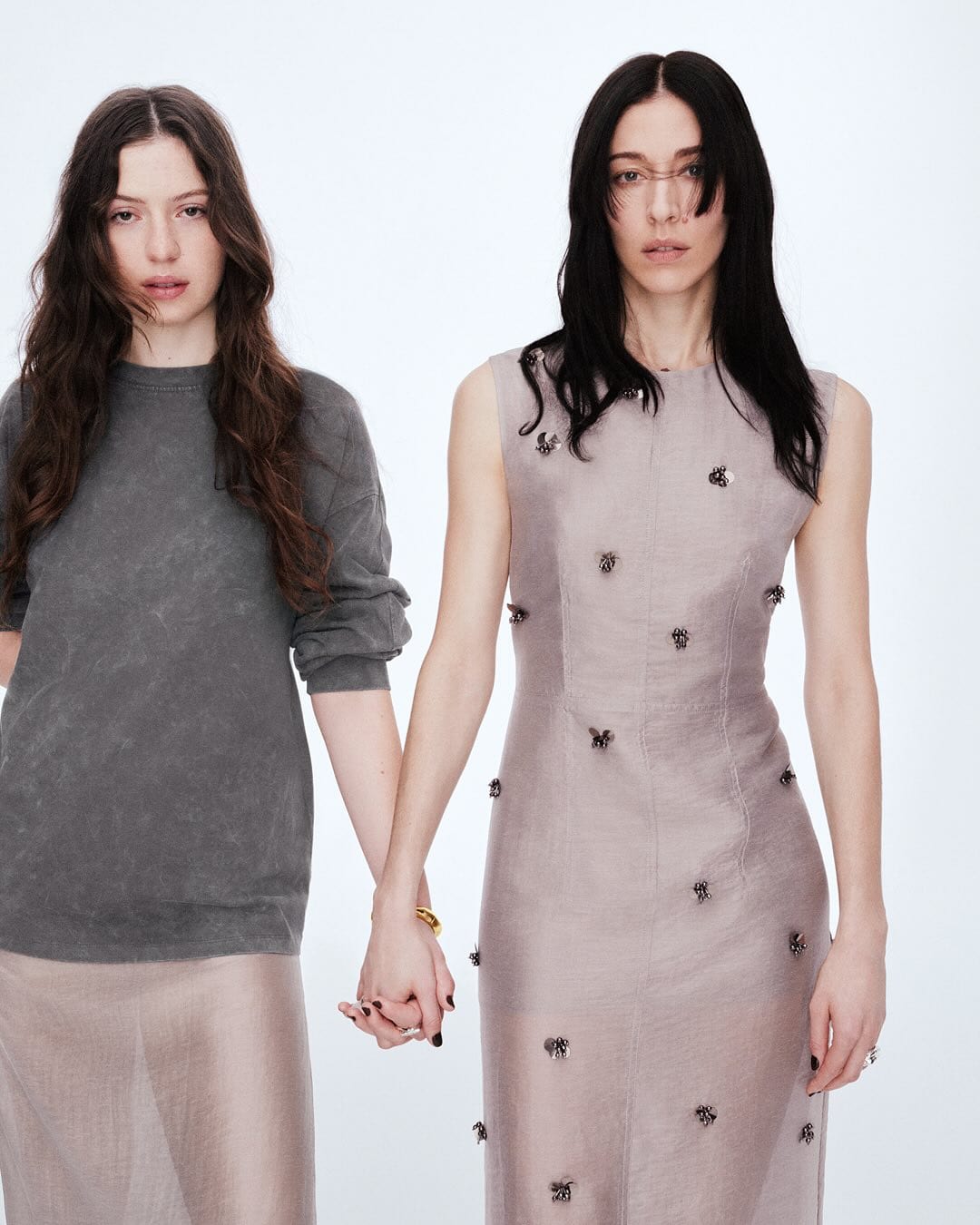 Two women holding hands against a white studio background wearing grey knitwear and a sleeveless embellished dress