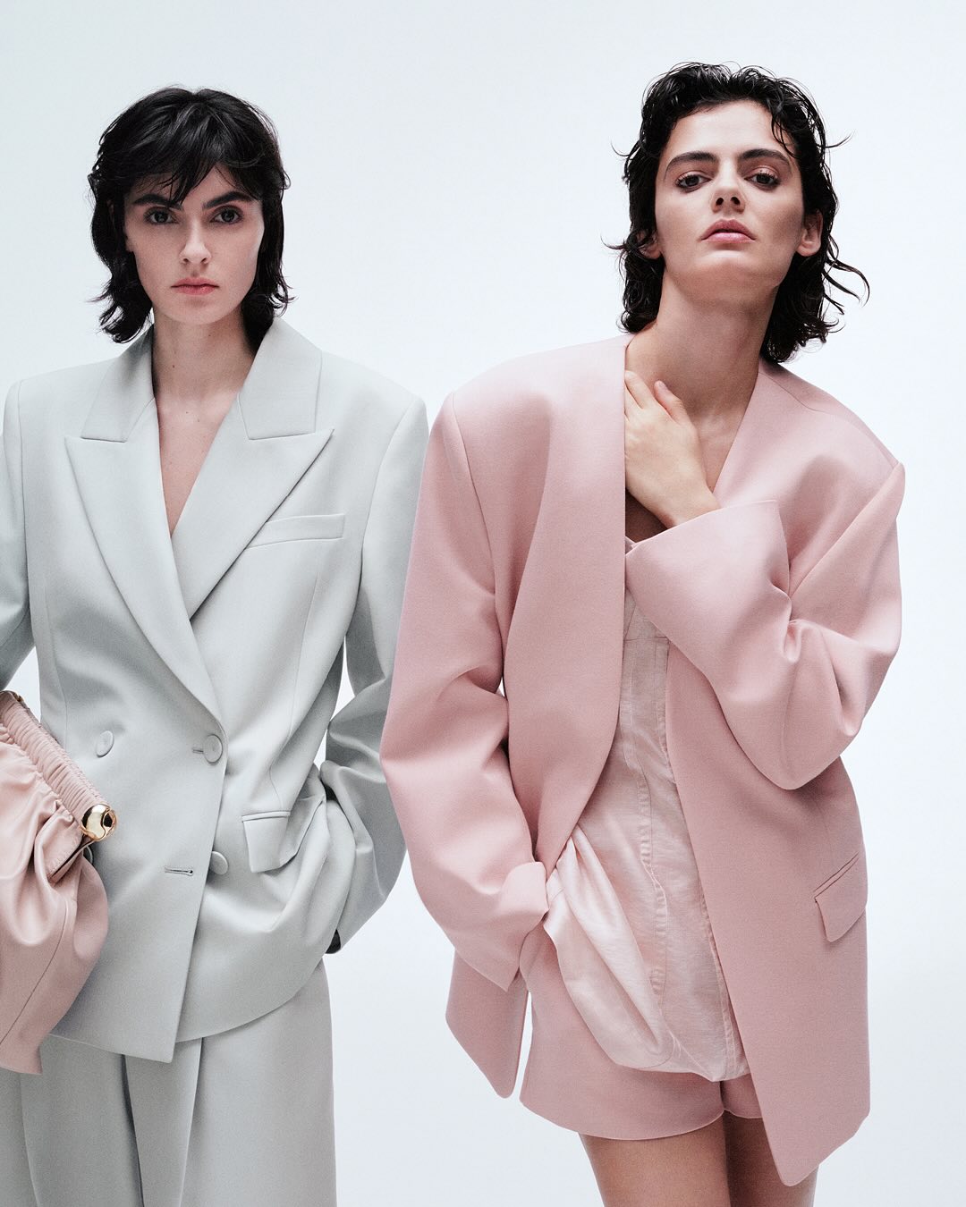 Two models wearing tailored oversized blazers in pale sage green and light pink against a seamless white background.
