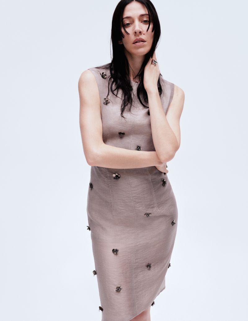 Caroline Polachek wearing a sleeveless taupe dress with dark embellishments against a plain white studio background.
