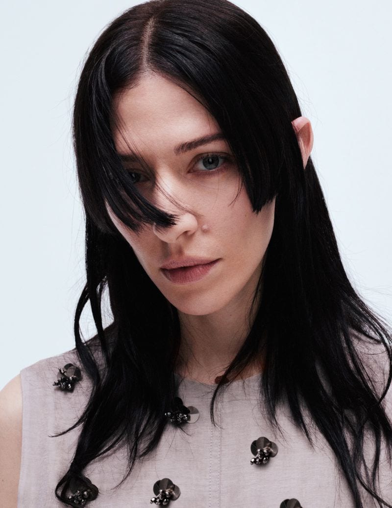 Portrait of Caroline Polachek with long black hair wearing a pale beige sleeveless top against a white studio background.