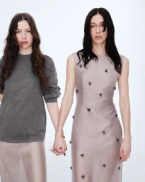 Two women holding hands against a white studio background wearing grey knitwear and a sleeveless embellished dress