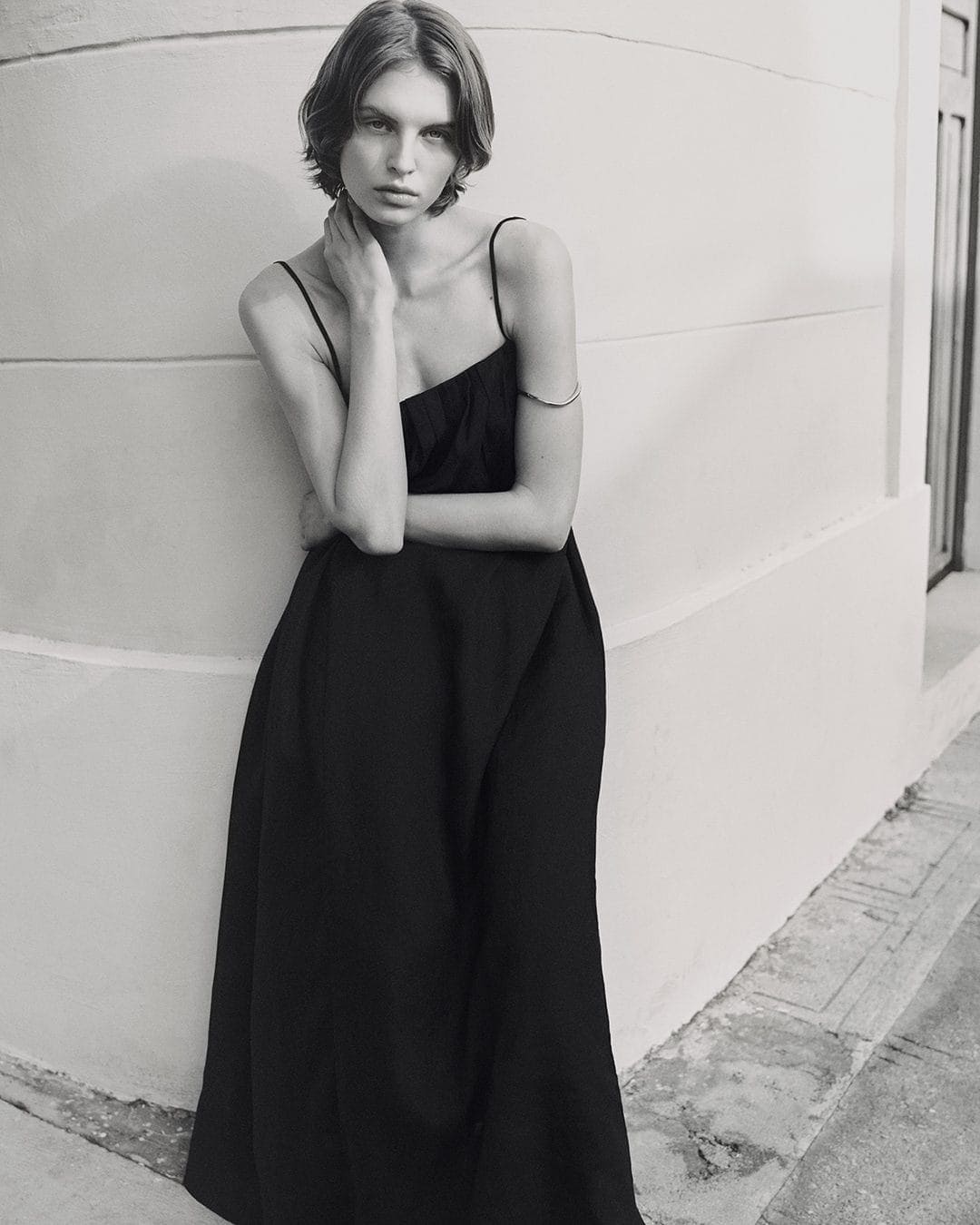 Model in a black slip dress leaning against a curved architectural wall in a monochrome fashion portrait.