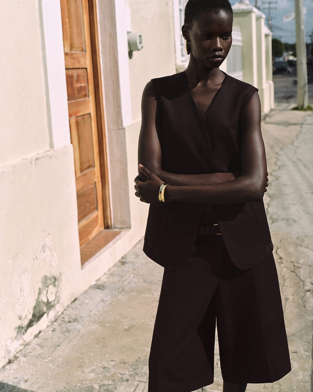 Model standing outdoors wearing a dark vest top and matching shorts against a pale building wall.