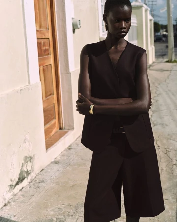 Model standing outdoors wearing a dark vest top and matching shorts against a pale building wall.