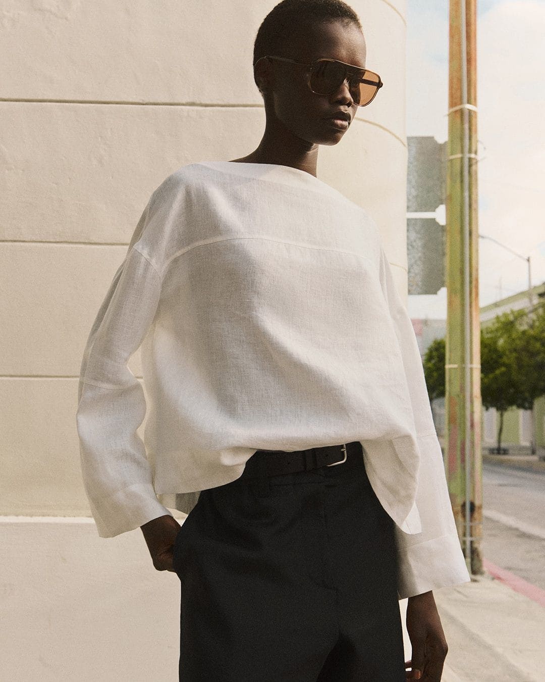 Model in oversized white top and black trousers wearing sunglasses outdoors against a building exterior wall