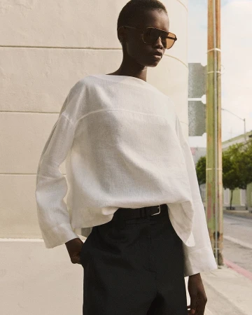 Model in oversized white top and black trousers wearing sunglasses outdoors against a building exterior wall