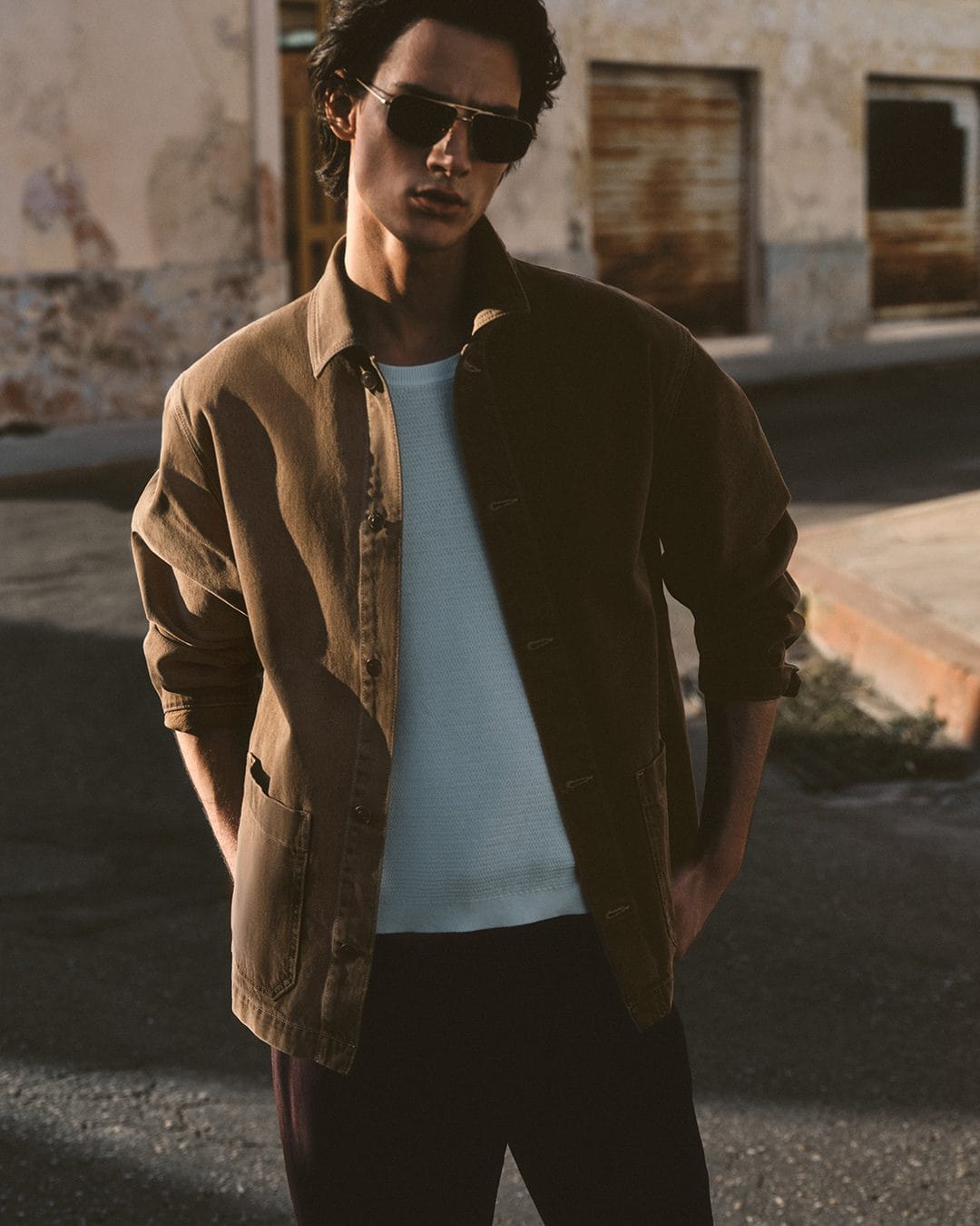 Man wearing sunglasses and a brown jacket standing outdoors on a sunny street for the COS Summer In The City campaign.