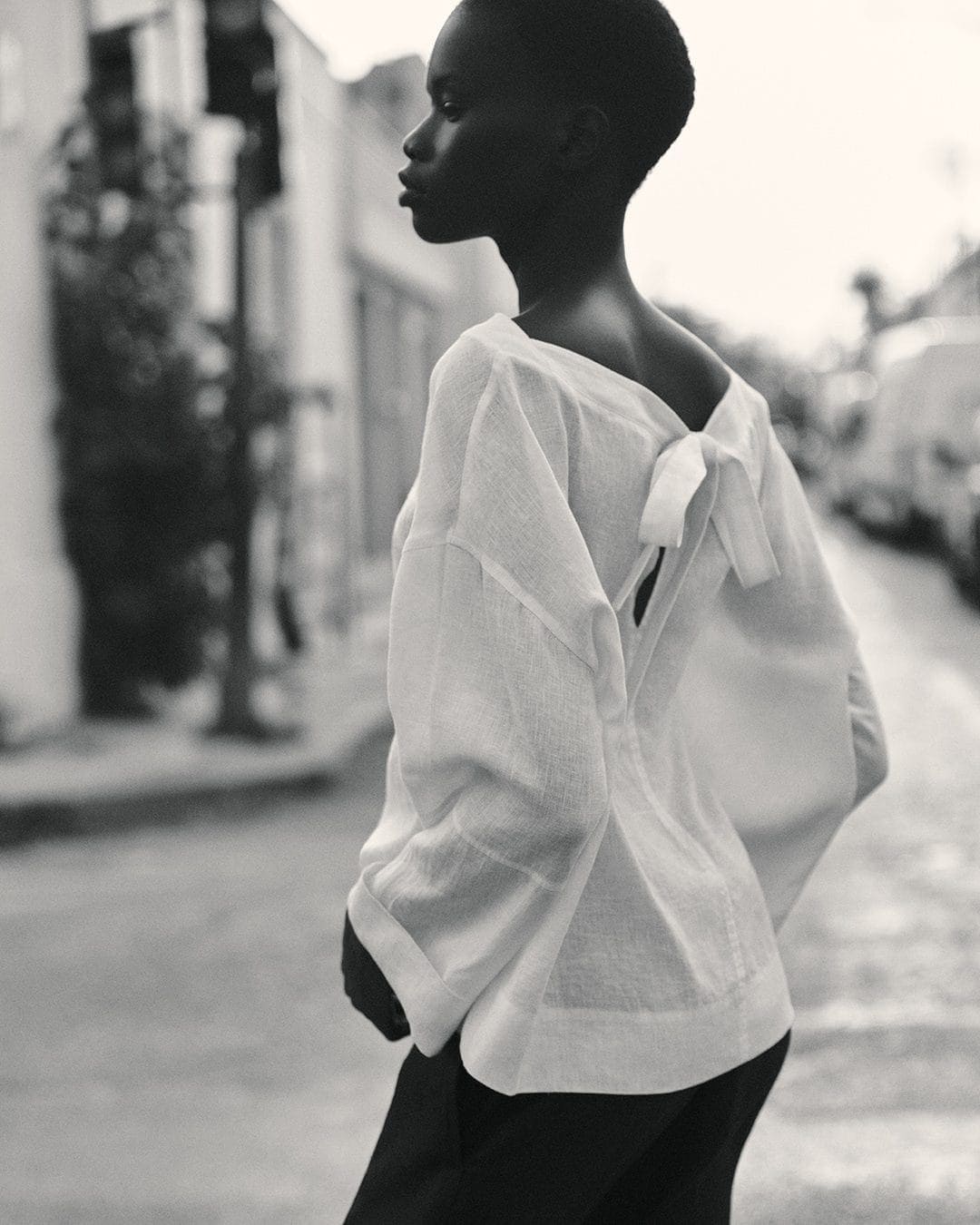 Model in profile wearing a white tied-back top and dark trousers on a city street