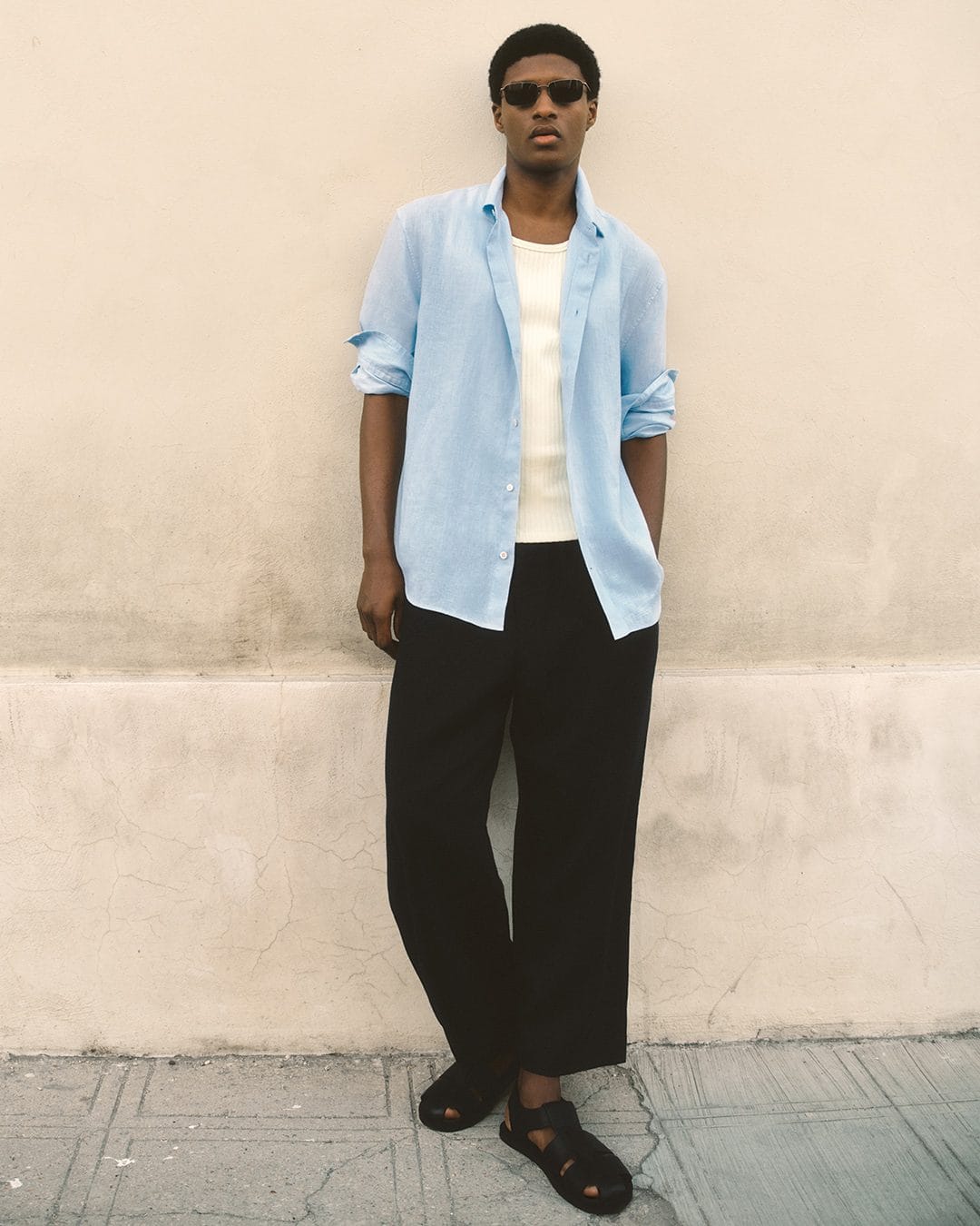 Man wearing sunglasses, unbuttoned light blue shirt, white tank top, and black trousers against a neutral wall