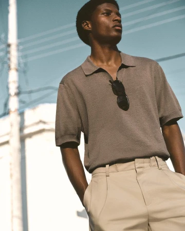 A man wearing a grey polo shirt with sunglasses tucked in, against a bright urban background.