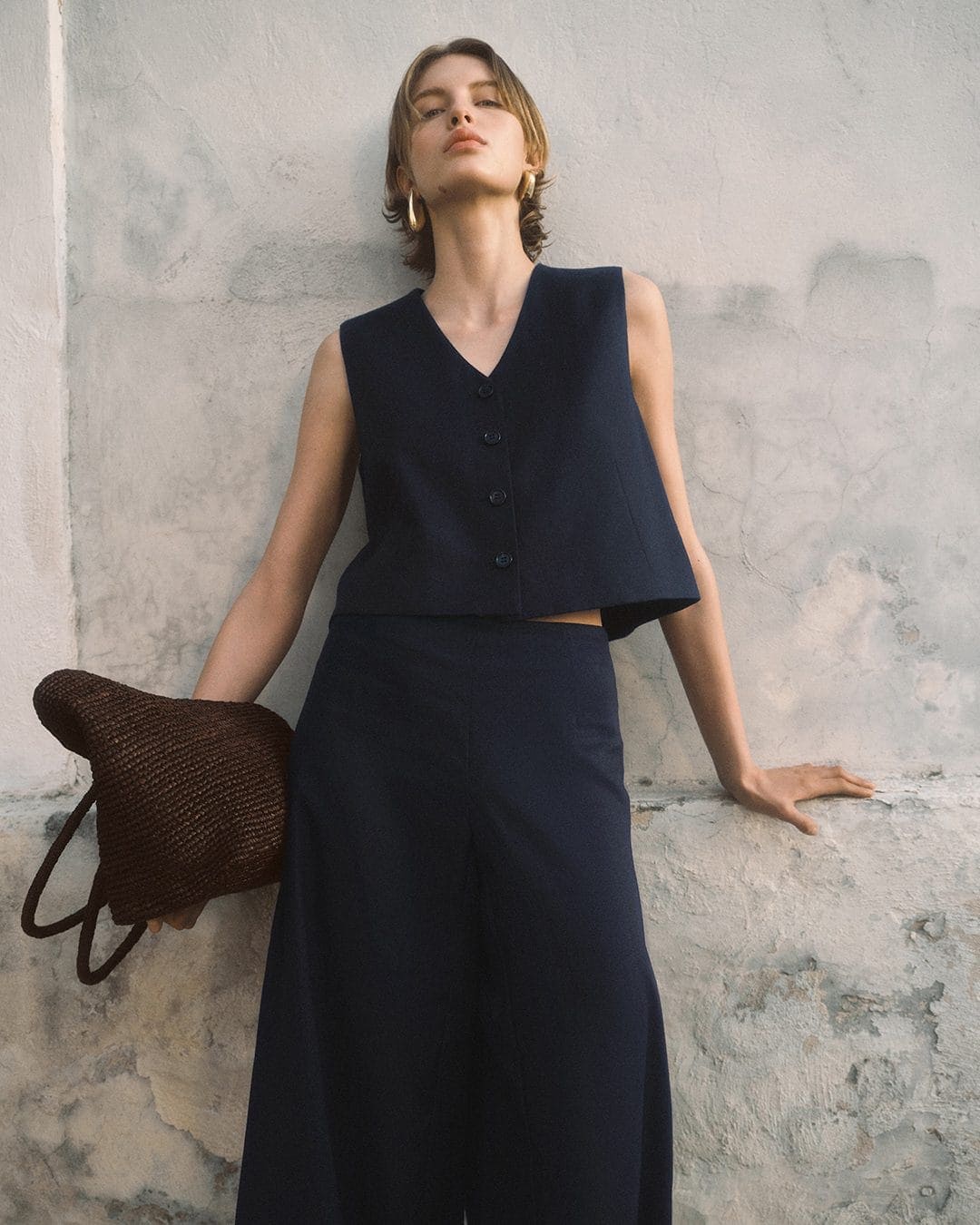 Model in navy vest and trousers holding a brown woven bag leaning against a weathered stone wall