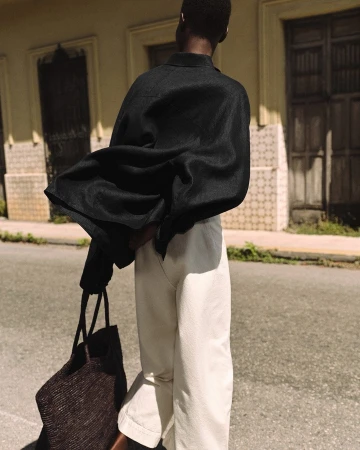 Model standing outdoors wearing a black buttoned shirt and white trousers holding a dark woven tote bag against an urban street backdrop