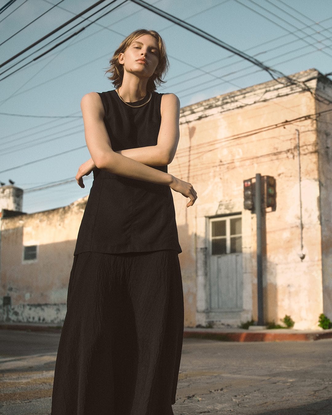 Model standing outdoors wearing a black sleeveless top and matching skirt under warm sunlight against a faded urban building