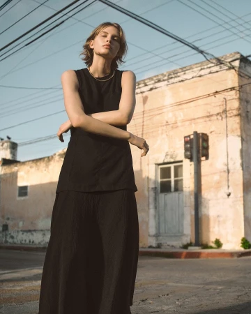 Model standing outdoors wearing a black sleeveless top and matching skirt under warm sunlight against a faded urban building