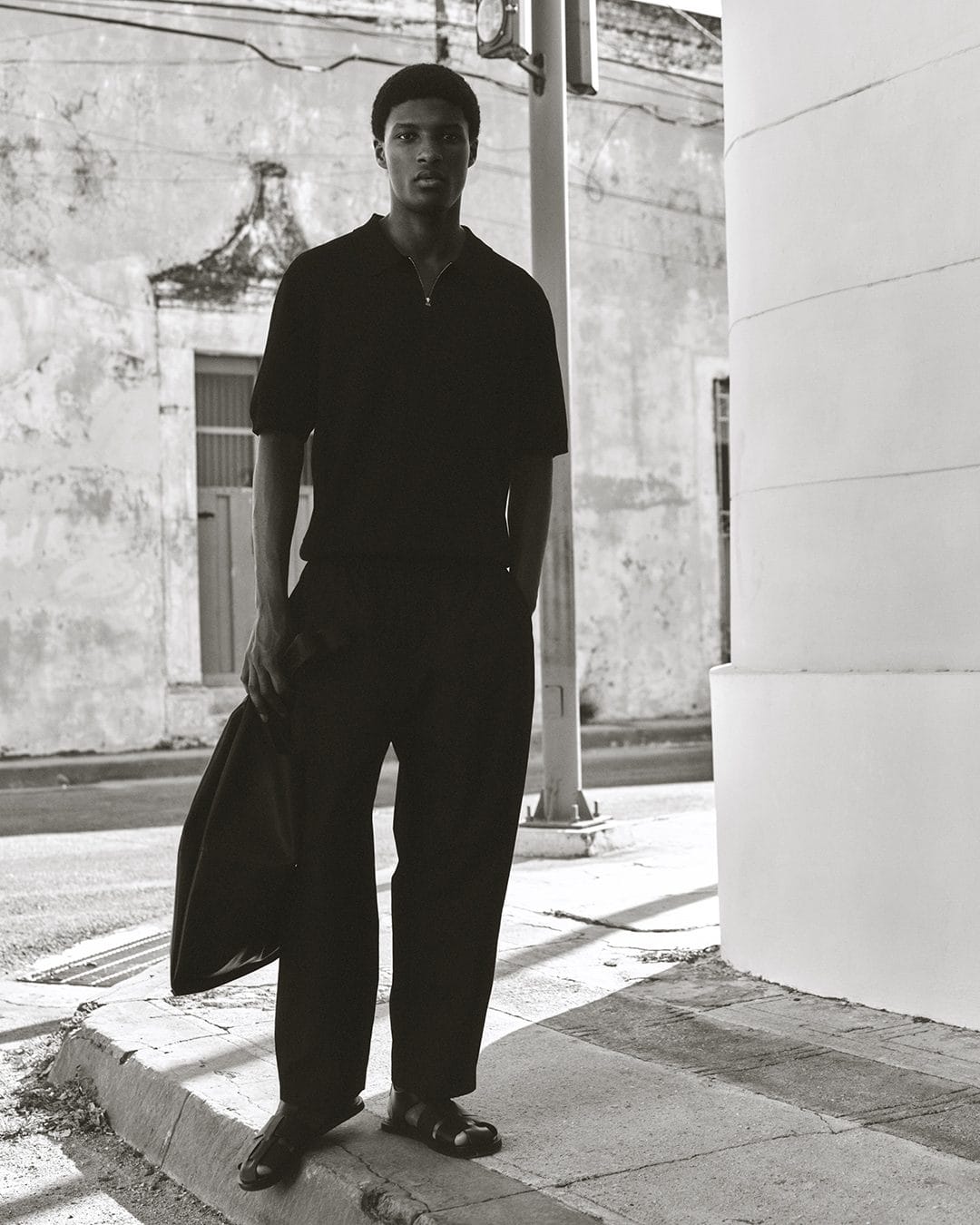 A black man wearing a dark polo shirt and trousers stands on a city street holding a bag in a monochromatic campaign image for COS.
