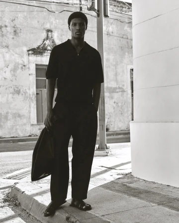 A black man wearing a dark polo shirt and trousers stands on a city street holding a bag in a monochromatic campaign image for COS.