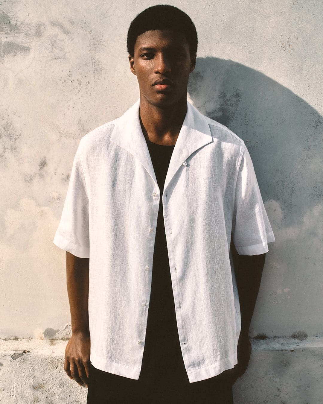 Young Black man wearing an unbuttoned white short-sleeve shirt against a textured concrete wall.