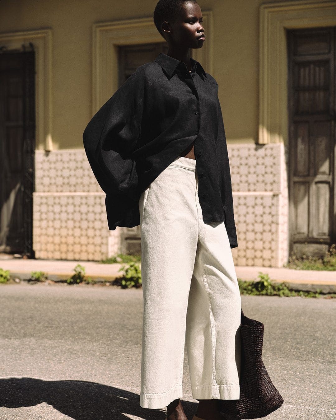 Model wearing a black button-down shirt and cream trousers standing on a sunny street for COS summer campaign.
