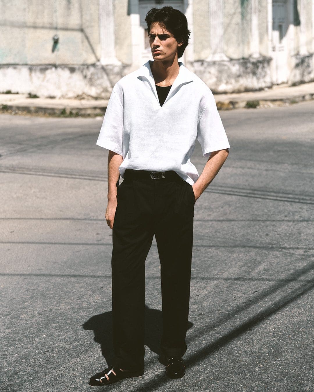 Man wearing a white knit polo shirt and black trousers standing outdoors on a paved street against a building wall