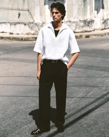 Man wearing a white knit polo shirt and black trousers standing outdoors on a paved street against a building wall