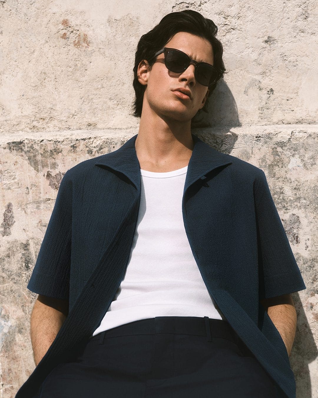Man wearing dark sunglasses, dark blue textured short-sleeve shirt, white undershirt, and dark trousers leaning against a concrete wall.