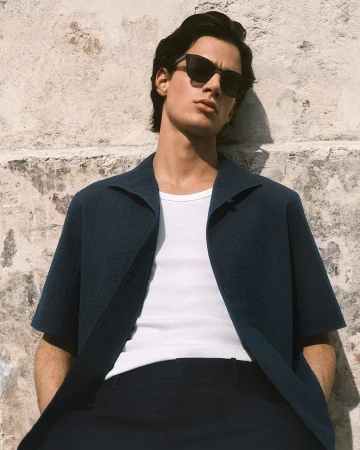 Man wearing dark sunglasses, dark blue textured short-sleeve shirt, white undershirt, and dark trousers leaning against a concrete wall.
