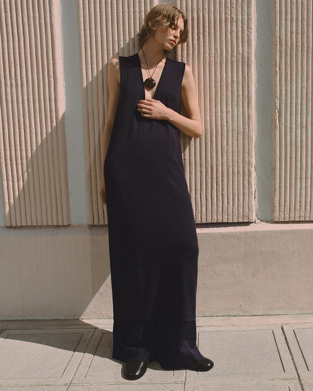 Model in a long black sleeveless dress leaning against a fluted concrete wall outdoors for the COS Summer In The City campaign.