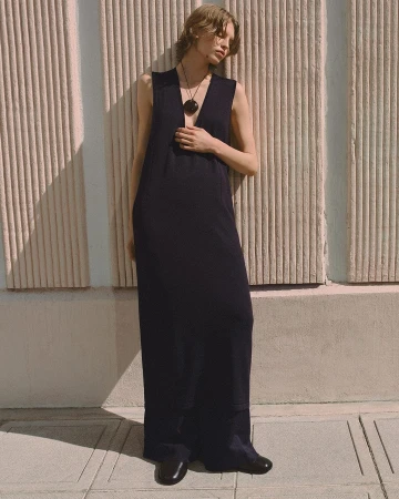 Model in a long black sleeveless dress leaning against a fluted concrete wall outdoors for the COS Summer In The City campaign.