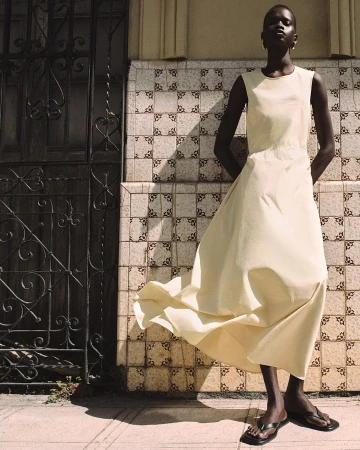 Model standing outdoors wearing a long cream sleeveless dress and black sandals against a patterned tile wall