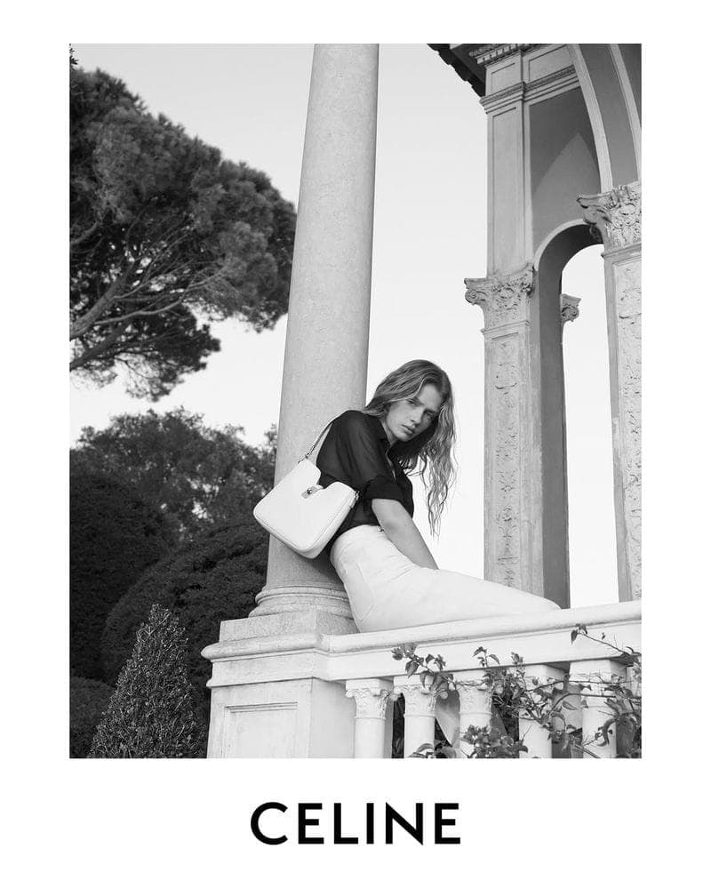 Model seated beside a stone pillar holding a white handbag for Celine Ete 2025 campaign