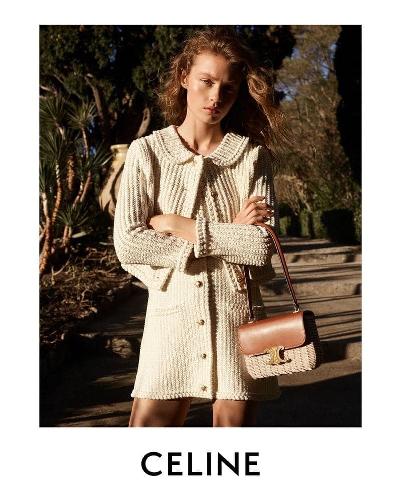 Model wearing a cream knit cardigan and skirt holding a structured brown leather handbag outdoors.