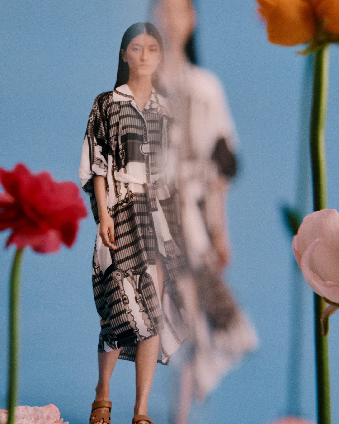 Model in printed patterned tunic dress standing against a blue background framed by foreground flowers