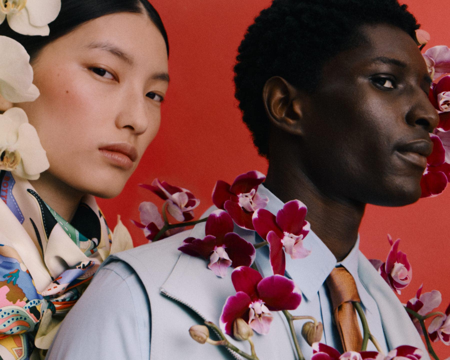 Close-up portrait of an East Asian woman with white orchids and a Black man with purple orchids against a red background.