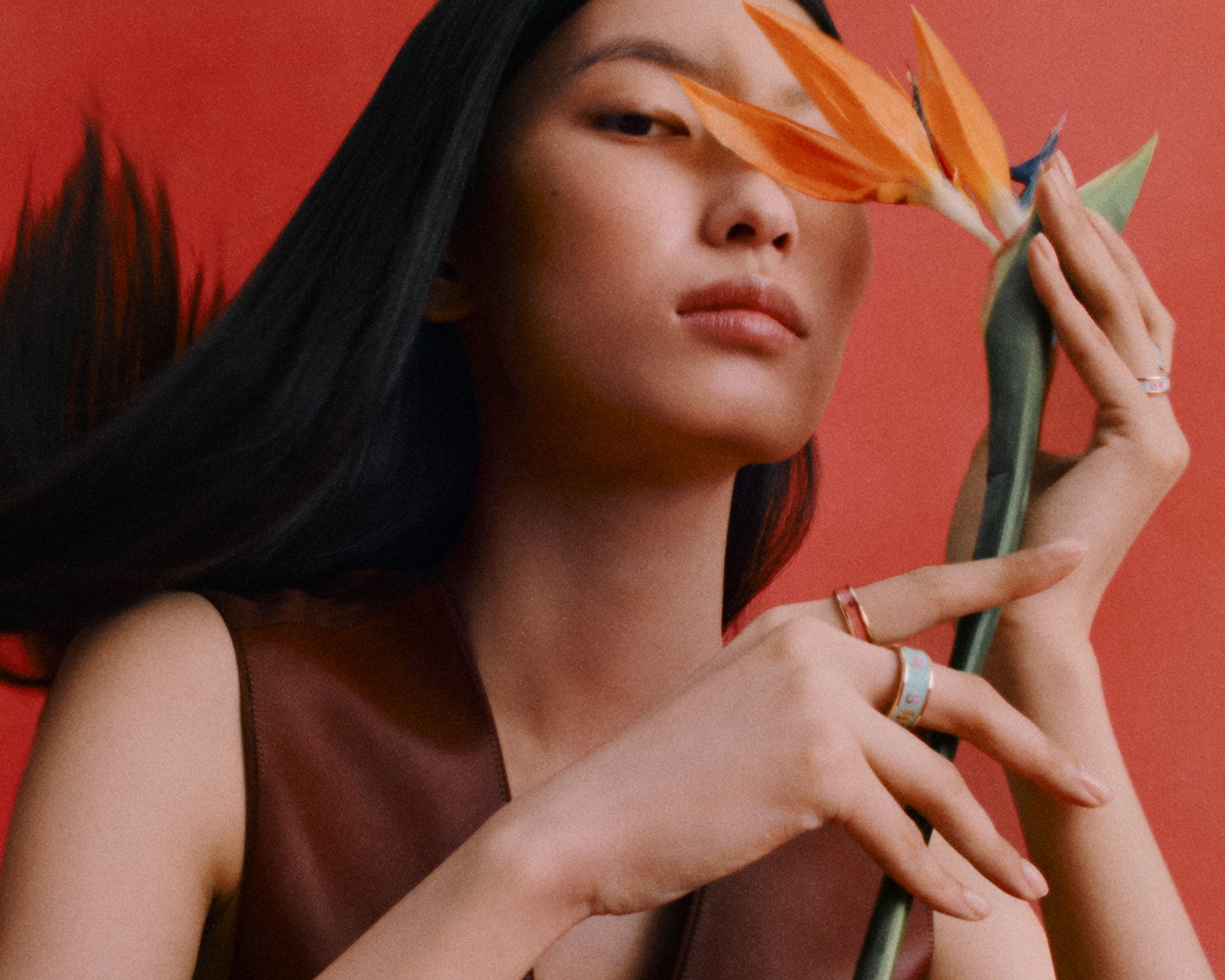 Model holding a bird of paradise flower near her face, wearing rings and a brown top against a red backdrop