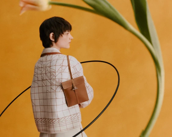 Model wearing a patterned jacket carrying a brown leather bag against a solid yellow backdrop