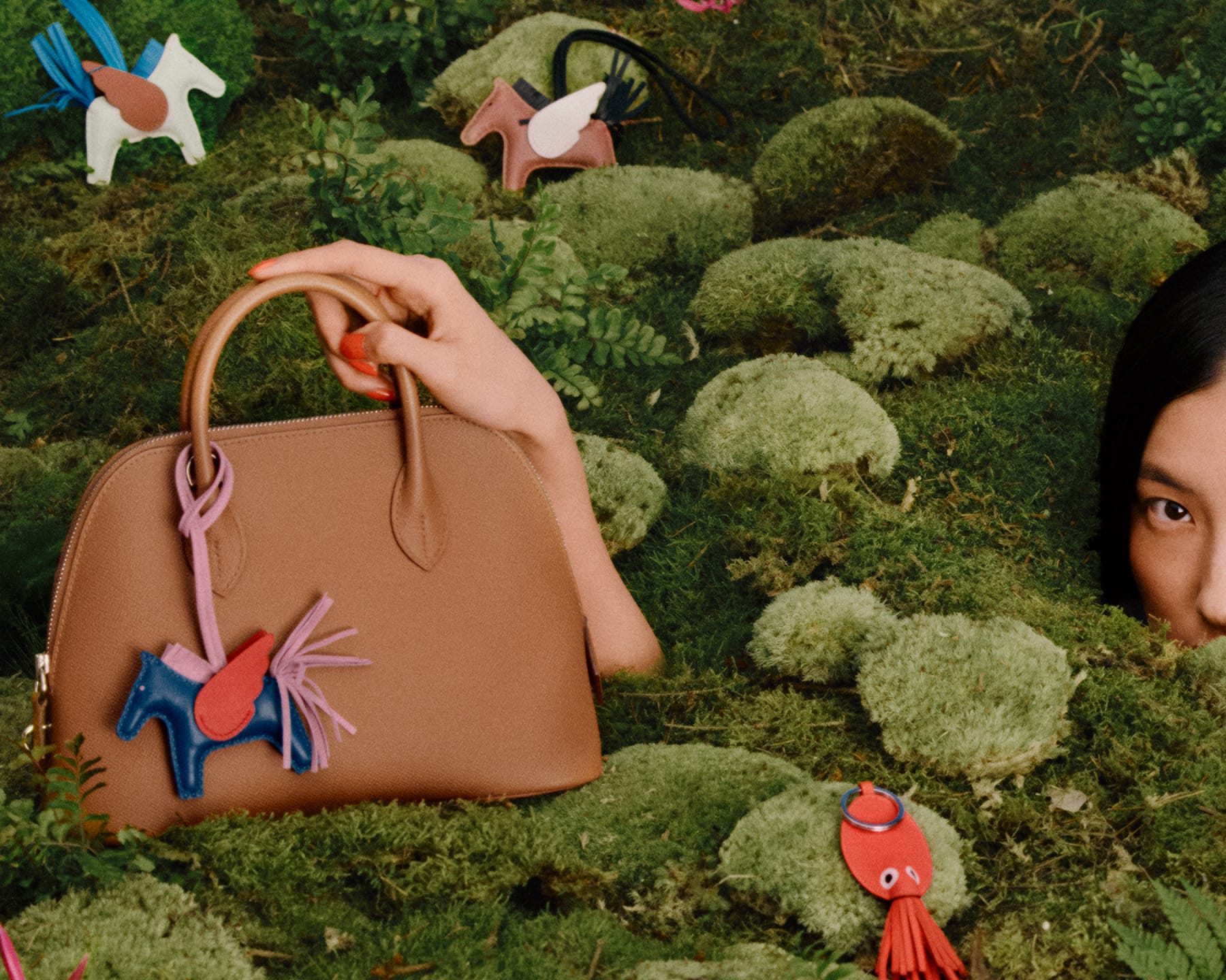 Tan leather handbag on a mossy green ground with whimsical winged horse bag charms in an outdoor nature setting