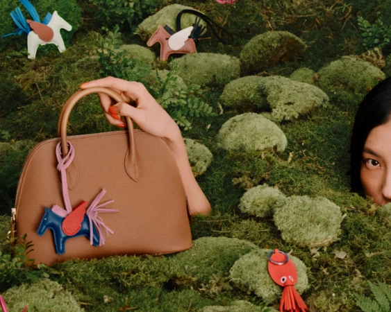 Tan leather handbag on a mossy green ground with whimsical winged horse bag charms in an outdoor nature setting
