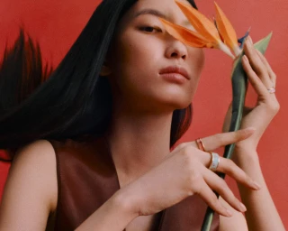 Model holding a bird of paradise flower near her face, wearing rings and a brown top against a red backdrop