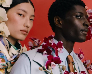 Close-up portrait of an East Asian woman with white orchids and a Black man with purple orchids against a red background.