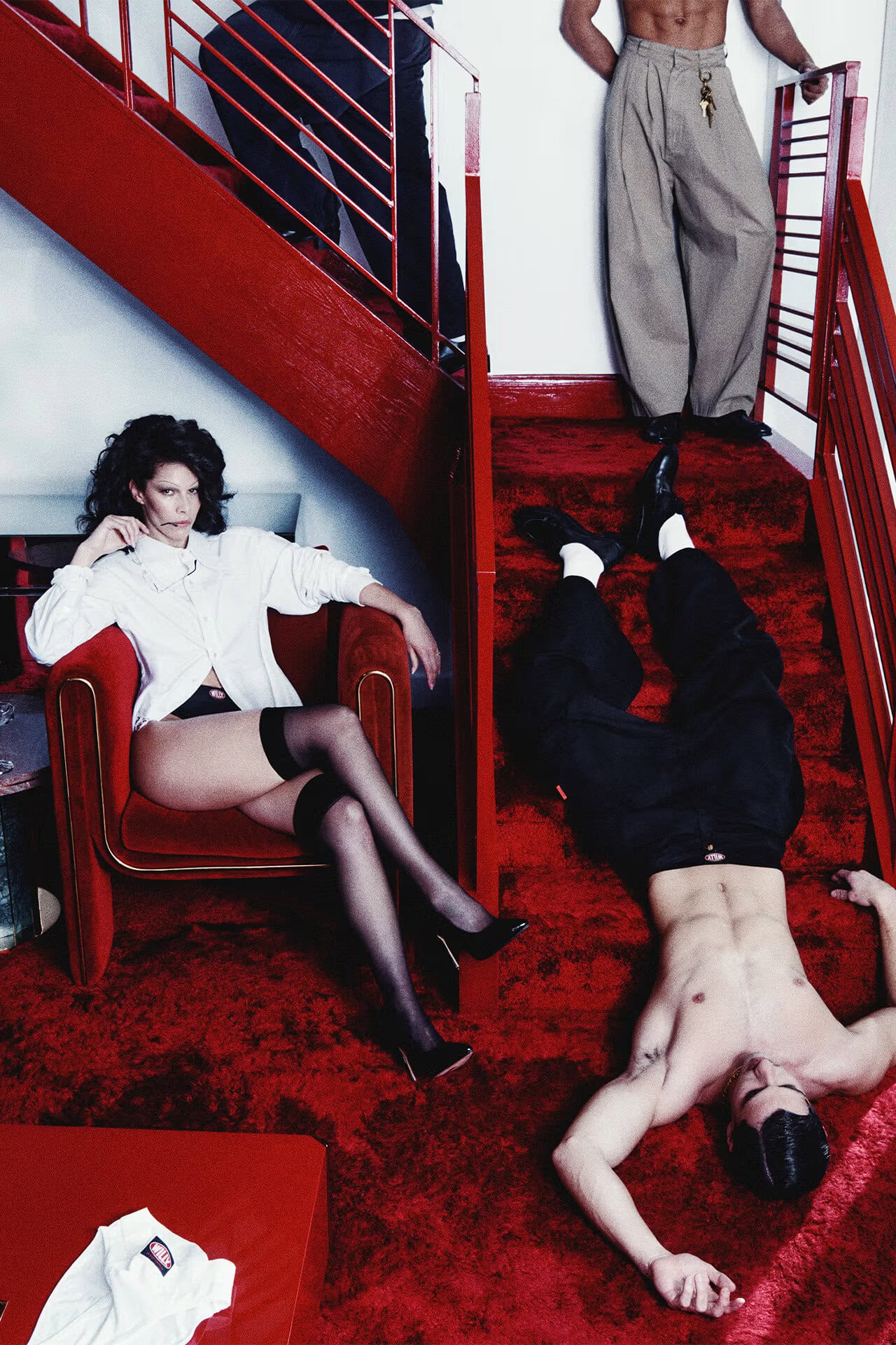 Model in white shirt and black stockings seated in a red chair next to a staircase covered in red carpet with a shirtless man lying on steps