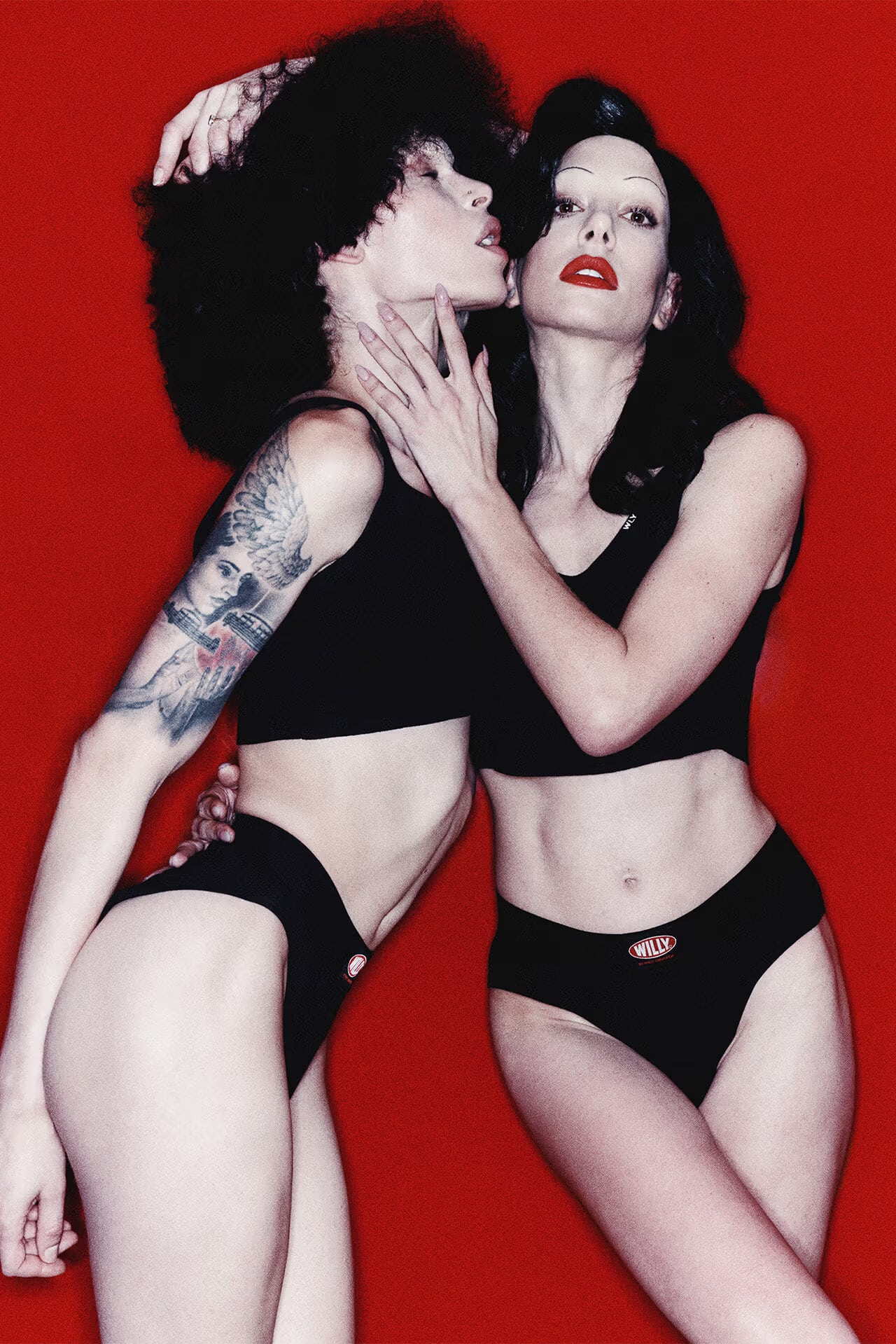 Two female models in black underwear posing against a vibrant red background for Willy Chavarria
